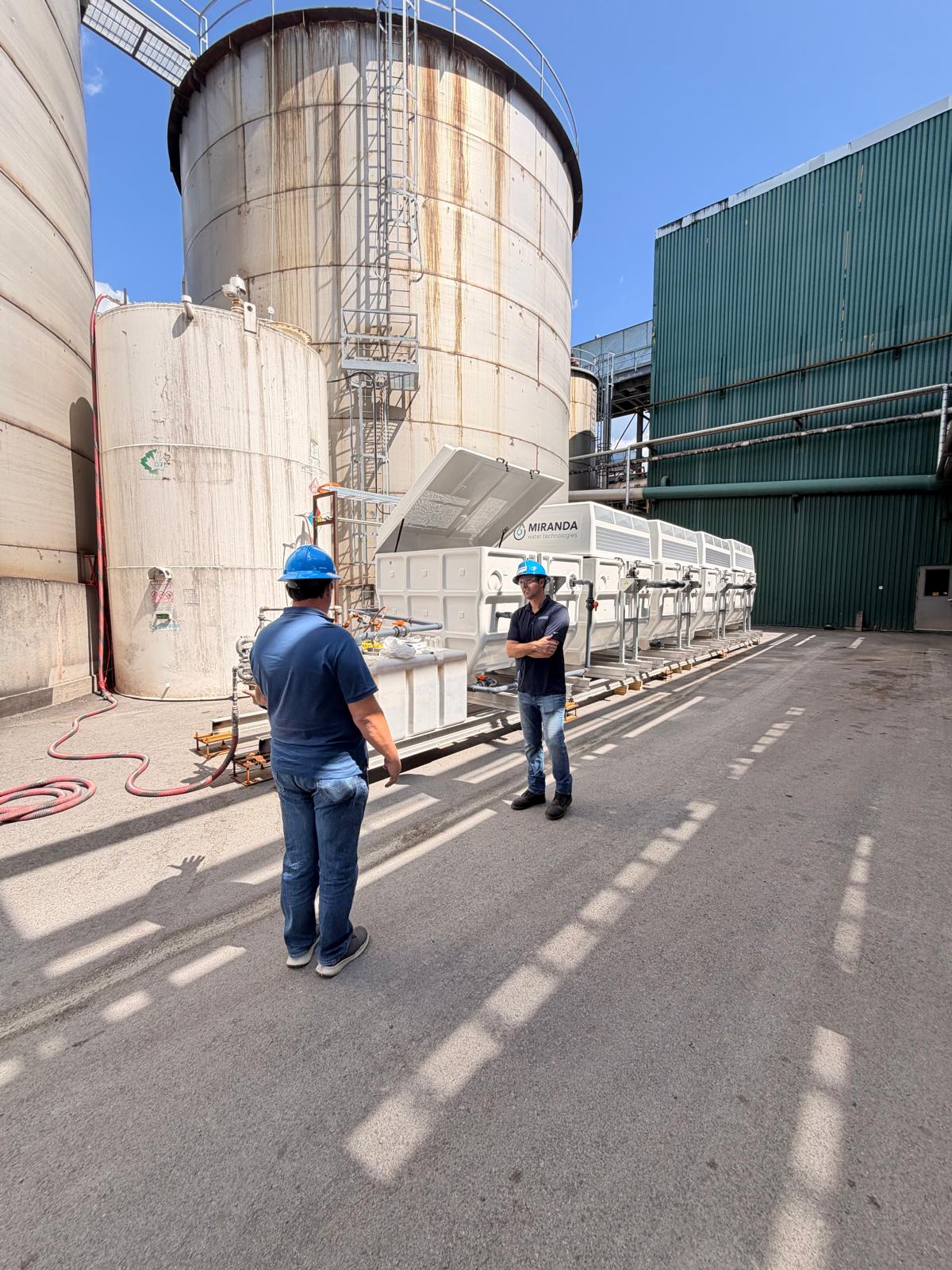 Two people in hard hats talking beside a row of white Miranda Miracell treatment modules installed at an industrial site, with storage tanks behind.