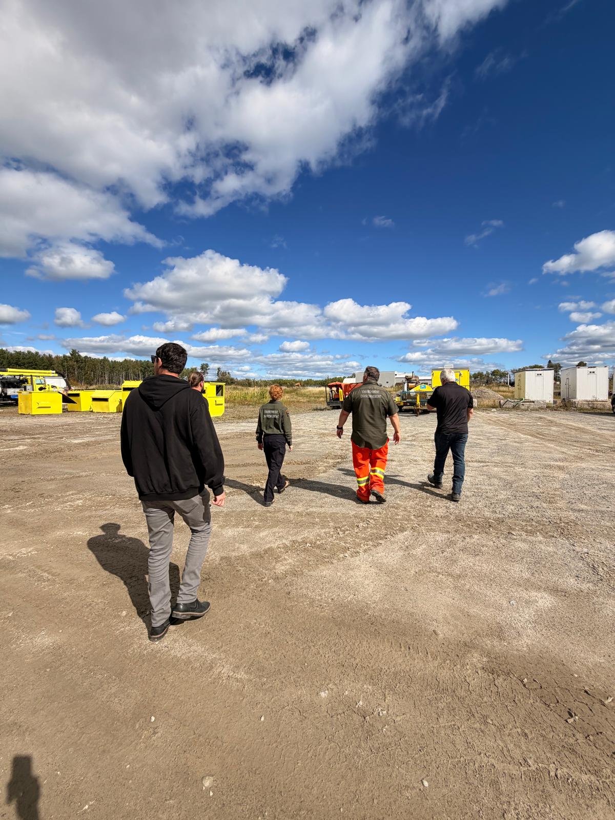 Five people, some in Environment-branded work shirts and high-visibility pants, walking across a gravel yard with yellow waste bins and site trailers under a blue sky.